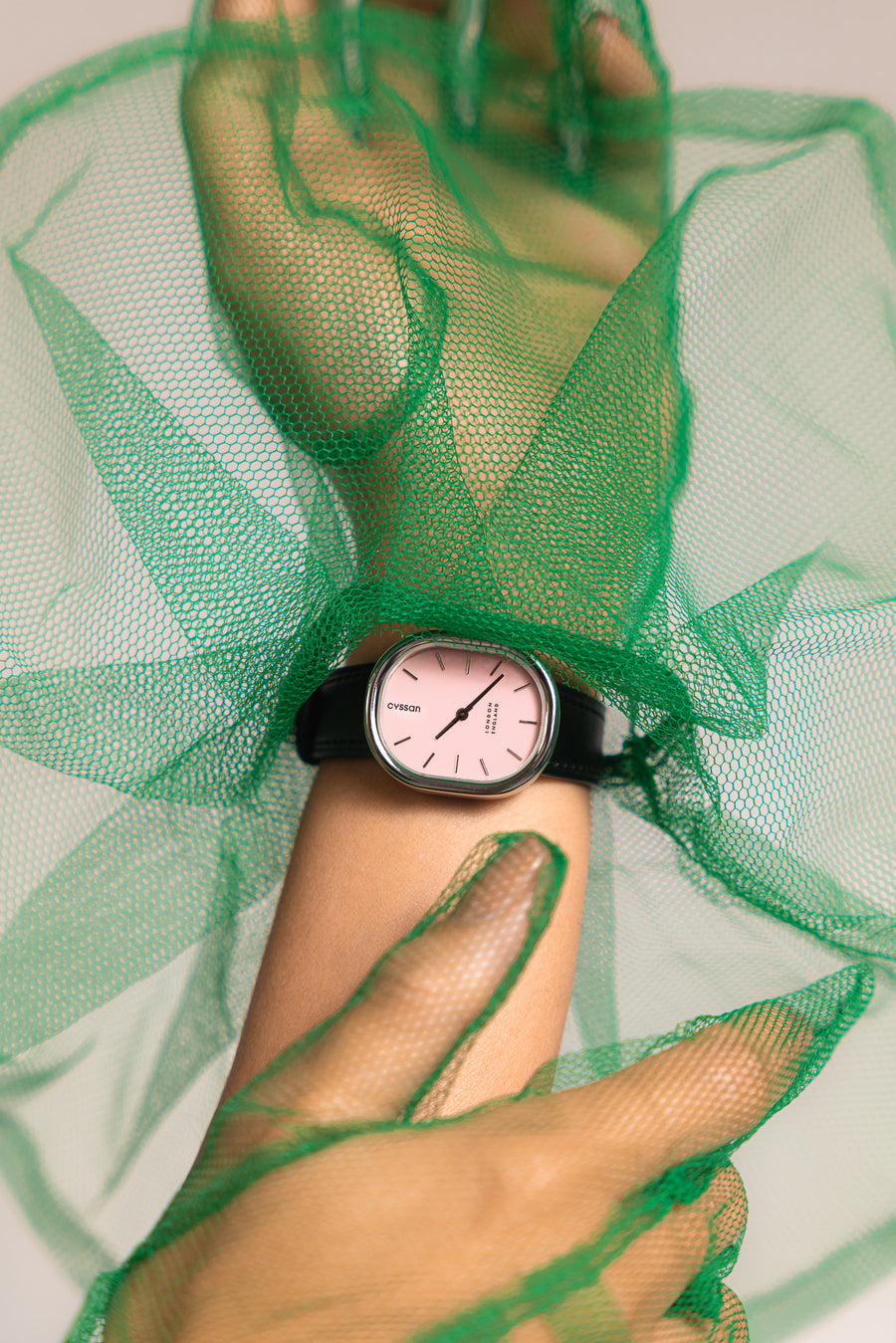 a close-up of a woman's hand and wrist. She's wearing a watch with a pink dial, silver case and black strap, and green tulle gloves