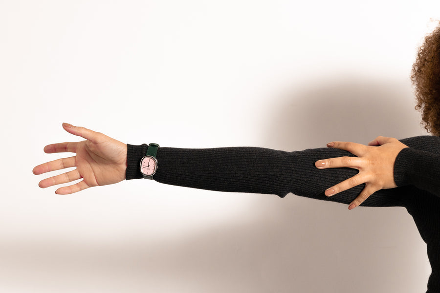 A woman's outstretched arm. She's wearing a dark grey long-sleeved dress with a wristwatch over the sleeve. The watch has a pink dial, a green strap and a silver case.