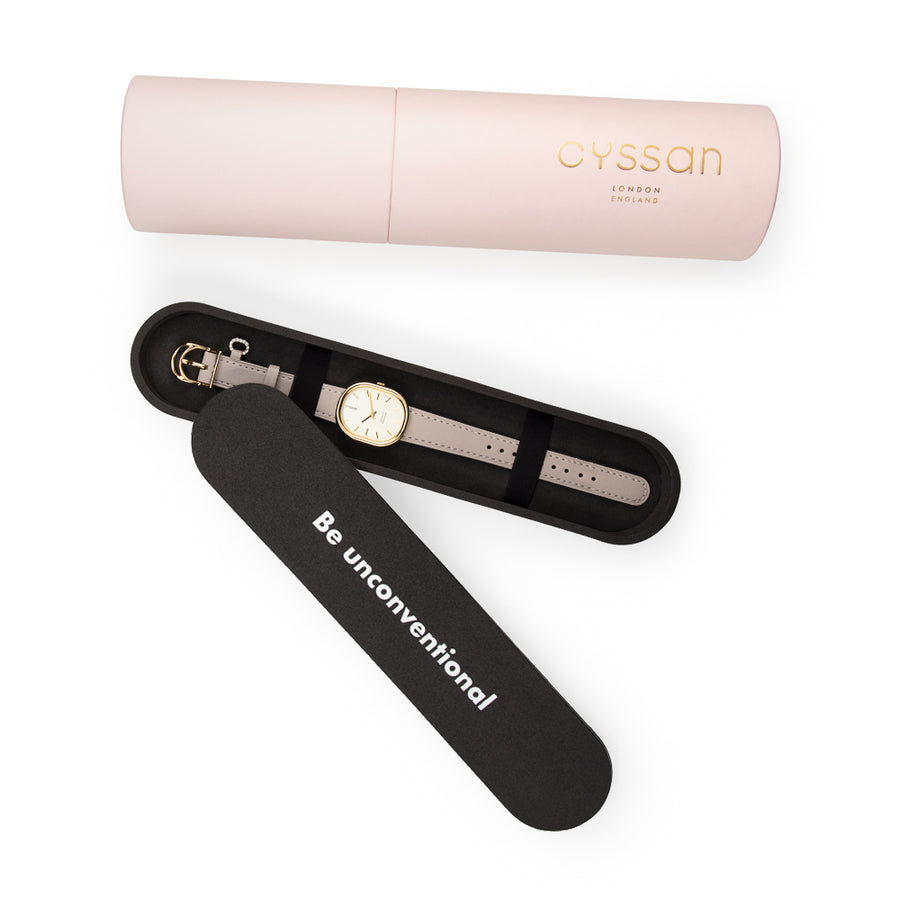 the Cyssan pink cylindric packaging. The black-foam inner packaging is open and there is a watch with a gold-plated watch case and sand-coloured watch strap placed inside. It is held in place with two black elastic straps.